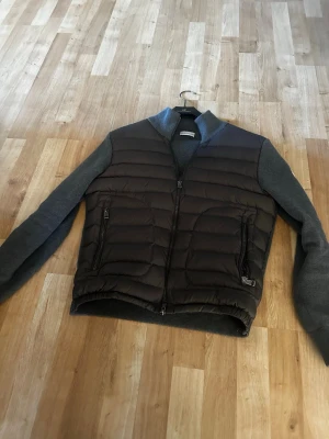Moncler cardigan  - Snygg dunväst från Moncler i mörkgrått med quiltad front och stickade ärmar. Västen har två dragkedjeförsedda fickor och ribbade muddar. Perfekt för lager-på-lager och ger en sportig men stilren look.