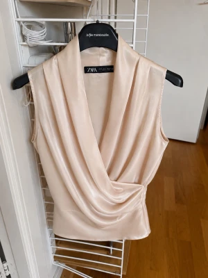 Champagnefärgad omlottopp från Zara - Säljer en champagnefärgad omlottopp från Zara i glansigt material. Toppen är ärmlös med draperad omlottfront och har en elegant, mjuk siluett. Perfekt att styla med jeans eller kjol för en trendig look.