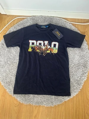  Polo t-shirt - Snygg t-shirt med stort färgglatt POLO-tryck och motiv av polospelare på bröstet. Klassisk rund hals och korta ärmar. Perfekt för dig som vill ha en ikonisk och sportig look.