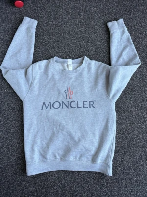 Grå sweatshirt från Moncler - Snygg grå sweatshirt från Moncler med rund halsringning och tryckt logga framtill i mörkblått och rött. Tröjan har långa ärmar och ribbade muddar vid ärmslut och nederkant. Perfekt för en avslappnad och trendig look.