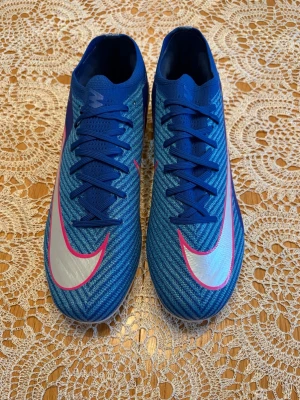 Blå Nike Mercurial fotbollsskor - Säljer ett par blå Nike Mercurial fotbollsskor med silverfärgade och rosa detaljer. Skorna har snörning, låg profil och är tillverkade i ett syntetiskt material med textilstruktur för bra bollkontroll. Perfekta för dig som vill ha stilrena och snabba skor på planen.