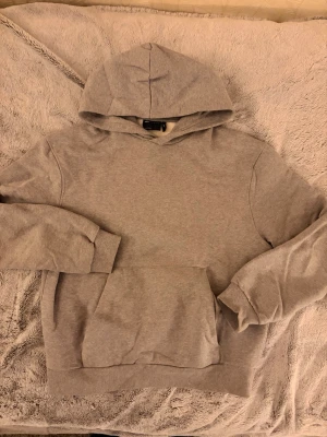 Grå hoodie från ASOS Design - En basic grå hoodie från ASOS Design med stor huva och magficka. Tillverkad i mjukt bomullsmaterial och har ribbade muddar vid ärmslut och nederkant. Perfekt för en chill och avslappnad stil.