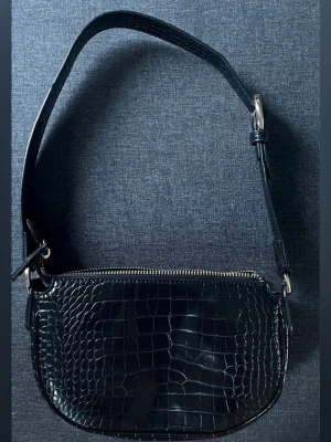 MING crocodile handväska  - Snygg svart handväska med axelrem i krokopräglat konstläder. Väskan har rundad form, justerbar rem med guldfärgat spänne och dragkedja upptill. Perfekt för dig som gillar trendiga accessoarer med lite edge.