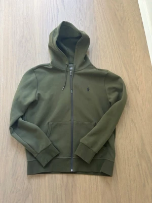 Olivgrön hoodie från Polo Ralph Lauren - Snygg olivgrön hoodie med dragkedja från Polo Ralph Lauren. Klassisk design med huva och dragsko, två fickor framtill och diskret logga broderad på bröstet. Säljer tröjan då den inte passade mig och den är enbart testad. Tröjan är i storlek Medium. Hör av er vid frågor!