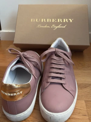 Rosa sneakers från Burberry - Säljer ett par snygga rosa sneakers från Burberry med vita sulor och gulddetalj vid hälen där Burberry-loggan syns. Skorna har klassisk låg modell, perforerade detaljer på sidorna och matchande rosa skosnören. Perfekta för dig som vill ha en stilren och exklusiv look.
