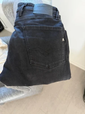 Svarta jeans från Replay Anbass  - Säljer ett par svarta jeans från Replay med klassisk femficksdesign och diskret logga på bakfickan. Jeansen har raka ben och normal midja, perfekta till en avslappnad stil. Materialet är mjukt och stretchigt för extra komfort.