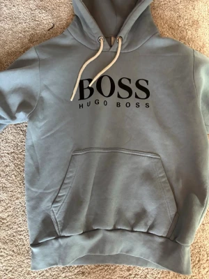 Grå hoodie från Hugo Boss - Snygg grå hoodie från Hugo Boss med stor svart BOSS-logga på bröstet. Hoodien har huva med vita dragsnören och en klassisk känguruficka framtill. Tillverkad i mjukt material som känns skönt mot huden. Perfekt för en avslappnad och trendig look.
