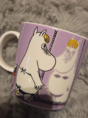 Lila Muminmugg med motiv - Säljer en lila mugg med Muminmotiv där Snorkfröken syns både framför en spegel och sittandes med presenter. Muggen är i keramik och har ett gulligt och färgglatt tryck som passar perfekt för Muminfans.