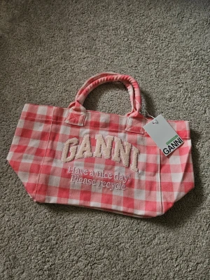 Rutig Tote Bag från Ganni - Supersöt handväska från Ganni i rosa och vitt rutigt tyg. Väskan har broderad text med 'GANNI' och 'Have a nice day! Please recycle' på framsidan. Två rejäla handtag och mjukt, slitstarkt material. Perfekt för att bära allt du behöver till skolan eller stan.