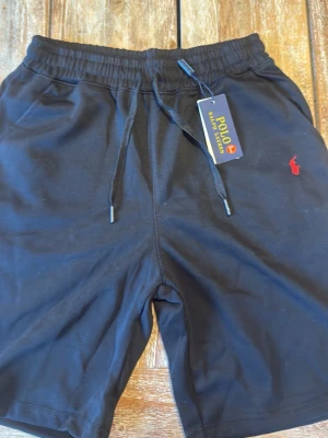 Mörk blåa shorts från Polo Ralph Lauren - Svarta shorts från Polo Ralph Lauren med dragsko i midjan och klassisk röd logga broderad på vänster sida. Tillverkade i mjuk bomull, perfekta för sommaren. Enkel och stilren design med normal passform.