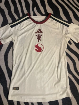 Manchester United away kit. - Snygg vit fotbollströja med Manchester United-logga och röd sponsorlogga på bröstet. Tröjan har mörkgröna ränder på axlarna, röda detaljer vid hals och ärmslut samt är tillverkad i ett lätt och ventilerande funktionsmaterial.