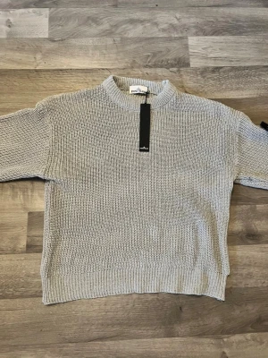 Ljusgrå stickad Stone Island tröja - En stilren ljusgrå stickad tröja med rund halsringning och ribbade muddar. Tröjan har en rak passform och är perfekt för lager-på-lager. Materialet ser ut att vara mjukt och bekvämt, vilket gör den enkel att matcha med olika outfits.