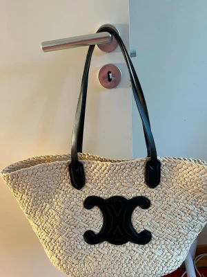 Beige stråväska med svarta detaljer - Snygg beige stråväska med svarta långa handtag och en stor svart dekorativ logga framtill. Väskan har en korgformad design och är tillverkad i flätat naturmaterial med detaljer i svart läderliknande material.