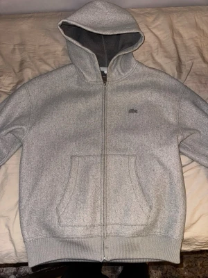 Lacoste tröja - En enkel och stilren grå hoodie med dragkedja framtill och stor huva. Hoodien har två rymliga fickor på magen och mjukt material på insidan. Perfekt för chill dagar eller när du vill ha en clean look. Ny pris 3000kr köpt i london 