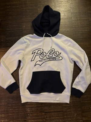 Vit och marinblå hoodie Polo Ralph Lauren - Snygg hoodie i vitt och marinblått från Polo Ralph Lauren. Stor logga framtill, kontrastfärgad huva och ficka på magen. Tillverkad i mjukt material, perfekt för en avslappnad och trendig look.