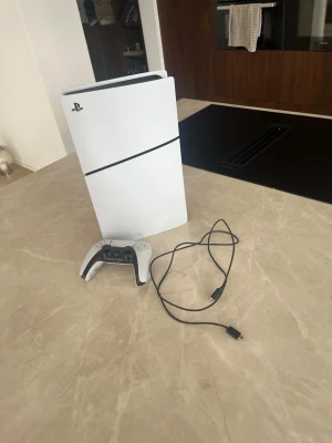 PlayStation 5 konsol med handkontroll - Säljer en vit PlayStation 5 konsol med tillhörande svartvit DualSense-handkontroll och USB-kabel. Konsolen har en futuristisk design med rundade kanter och matta paneler. Perfekt för gaming och streaming.