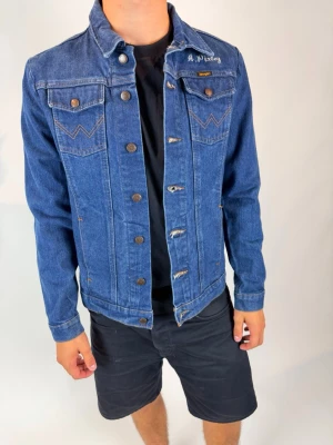 Wrangler Denim Jacket - This is a classic Wrangler blue denim jacket in size Medium. It features button closures, two chest pockets with button flaps, and signature 'W' stitching on the pockets. The jacket has a regular fit and is styled here with a black t-shirt and black shorts. The left chest area is embroidered with 'A. P. Lacey'.