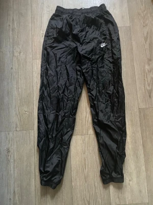 Svarta mjukisbyxor från Nike - Säljer ett par svarta mjukisbyxor från Nike med resår i midjan och anklarna. Byxorna har en avslappnad passform och är tillverkade i ett glansigt, lätt material. Nike-loggan syns tydligt på vänster ben. Perfekta för chill eller träning.
