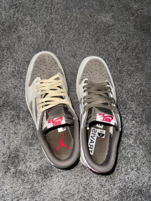 Nike Air Jordan 1 Low Travis Scott - Säljer ett par Nike Air Jordan 1 Low Travis Scott sneakers i brunt, vitt och beige med ikonisk omvänd Swoosh. Skorna har mockadetaljer, perforerad tå och olika färg på skosnörena. Röd logga på plösen och häl. Perfekt för dig som gillar streetwear.