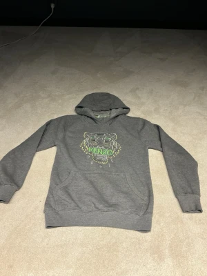 Grå Kenzo hoodie med tigertryck - Snygg grå hoodie från Kenzo med ikoniskt tigertryck och Kenzo Paris-logga i neonfärger på bröstet. Hoodien har huva och magficka, samt ribbade muddar vid ärmslut och nederkant. Perfekt för en cool och avslappnad stil.