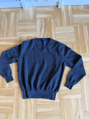 Mörkgrå stickad tröja med V-ringning - Mörkgrå stickad tröja från Polo Ralph Lauren med V-ringning och diskret röd logga broderad på bröstet. Tröjan har långa ärmar, ribbade muddar och nederkant. Perfekt för lager på lager och enkel att matcha med olika outfits.