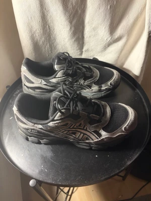 Svarta och grå sneakers från Asics - Säljer ett par svarta och grå sneakers från Asics med mesh- och mockadetaljer. Skorna har klassisk låg profil, snörning och tydliga Asics-loggor på sidorna. Perfekta för dig som gillar sportig och stilren look.