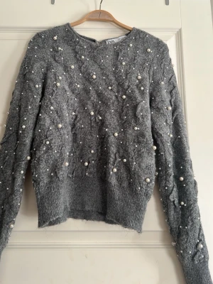 Grå stickad tröja med pärlor från Zara - Supersnygg grå stickad tröja från Zara med dekorativa pärlor över hela tröjan. Tröjan har rund hals, lång ärm och ribbade muddar. Perfekt för dig som vill ha något extra till din outfit. Materialet är mjukt och mysigt.