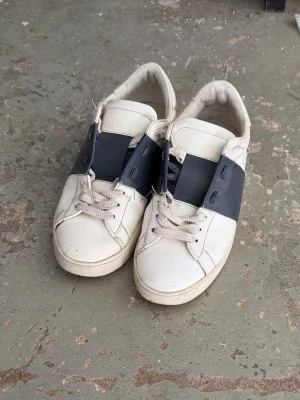 Vita sneakers med blå detalj - Säljer ett par vita sneakers med bred marinblå rem över sidan. Skorna har klassisk låg profil, vita snören och är tillverkade i slätt läder. Perfekta för dig som gillar stilrena och minimalistiska skor.