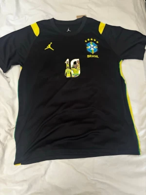 Svart Brasilien fotbollströja - Snygg svart fotbollströja med gula detaljer och Brasilien-emblem på bröstet. Nummer 10 och en bild på en spelare pryder framsidan. Tröjan har rund hals och korta ärmar, perfekt för träning eller match.