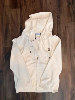 Vit hoodie med dragkedja från Moncler - Snygg vit hoodie från Moncler med dragkedja framtill och justerbar huva med snören. Klassisk Moncler-logga på bröstet och ribbade muddar vid ärmslut och nederkant. Perfekt för en clean och stilren look.