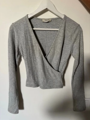 Grå omlottopp från Chiquelle - Säljer en ljusgrå ribbad omlottopp från Chiquelle i storlek XS. Toppen har lång ärm, croppad passform och djup v-ringning. Perfekt att styla med höga jeans eller kjol för en trendig look. Har ett litet hål som knappt syns, skriv för bild.