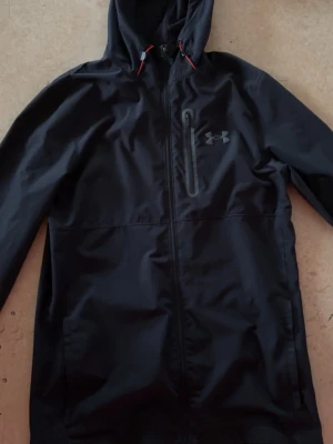 Svart vindjacka från Under Armour - Svart vindjacka från Under Armour med huva och röda dragskor. Sällsynt jacka då den inte säljs någonstans längre som t.ex jd sports eller under armour. Mycket bra skick. Kan endast frakta genom instabox!