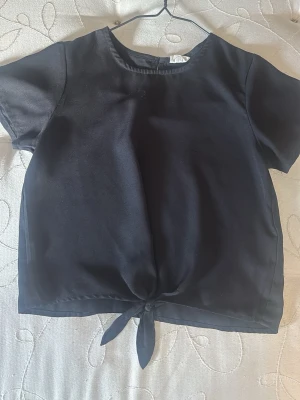 Svart kortärmad topp med knyt - Stilren svart topp i blus material med korta ärmar och rund halsringning. Nedtill finns en snygg knytning som ger en trendig touch. Perfekt att matcha med jeans eller kjol för en enkel men chic look.