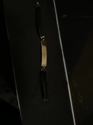Svart flätat armband från Guess - Snyggt armband från Guess med svart flätat band och en guldfärgad metallplatta där märket är ingraverat. Armbandet har stilrena detaljer och ett modernt, minimalistiskt uttryck. Perfekt accessoar för dig som gillar klassisk men trendig stil.