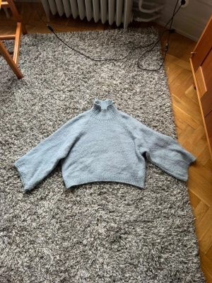 Ljusblå stickad trekvartsärmad polotröja - Mysig ljusblå stickad tröja med hög krage och vida ärmar. Tröjan har en boxig, avslappnad passform och är stickad i ett mjukt material som känns skönt mot huden. Har trekvartsärm.