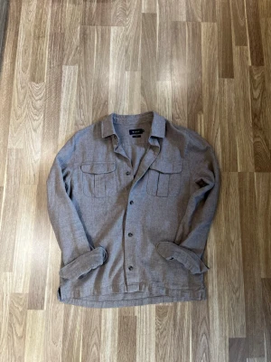 Riley Linne Overshirt - Stilren beige overshirt med knappar framtill och två stora bröstfickor med lock. Skjortkrage och långa ärmar. Perfekt lager-på-lager-plagg i ett mjukt tyg som ger en avslappnad look. Den är 100% linne. Sparsamt använd.