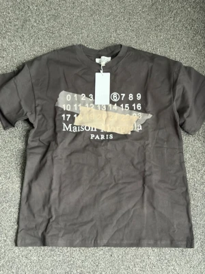  Maison Margiela t-shirt - T-shirt från Maison Margiela med ikoniskt tryck framtill och logga. Klassisk passform med rund hals och korta ärmar. Perfekt för dig som gillar streetwear och vill ha något stilrent men ändå unikt.
