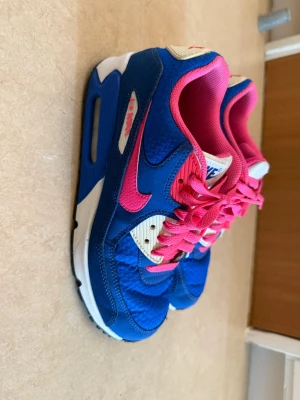 Nike Air Max 90 blå och rosa sneakers - Nike Air Max 90 sneakers i blått och rosa med vita detaljer. Skorna har en klassisk låg siluett, rosa snören och meshpartier för extra komfort. Swoosh-logga i rosa på sidan och vadderad insida i rosa. Perfekta för dig som vill sticka ut med färg. Strl 40, fint skick men som bilden visar lite gulnade/färgade i bak 