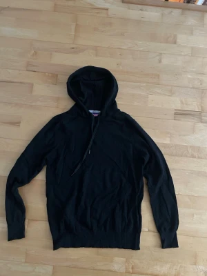 Svart kashmir hoodie med huva - En enkel svart hoodie med huva och dragsko. Hoodien har långa ärmar och ribbade muddar vid ärmslut och nederkant. Perfekt för en avslappnad och stilren look. Tröjan är 45% kashmir, 20% silke, 20% viskos och 15% elastane. Storlek M.