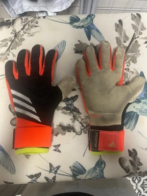 Adidas predator målvaktshadskar - Snygga målvaktshandskar från Adidas med URG 2.0-grepp. Handskarna är svart, orange och har vita detaljer samt breda kardborreband vid handleden. Perfekta för fotbollsmatcher och träning.