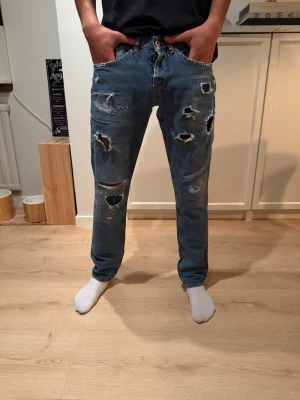 Dondup George  - Säljer ett par blå jeans med flera slitningar och hål på både fram- och baksida. Jeansen har en rak passform och klassisk femficksdesign. Perfekta för dig som gillar en edgy och avslappnad stil.