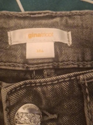 Grå jeans från Gina Tricot - Säljer ett par grå jeans från Gina Tricot i storlek 164. Jeansen har raka ben, klassiska bakfickor med knappdetalj och är tillverkade i ett mjukt denimtyg. Perfekta för en avslappnad och trendig look.
