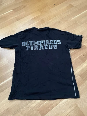 Svart Olympiacos Piraeus t-shirt,XL - Snygg svart t-shirt med Olympiacos Piraeus-tryck på ryggen och klubbmärke på bröstet. T-shirten har korta ärmar och vita ränder längs sidorna. Perfekt för dig som gillar sportig stil och vill visa ditt stöd för laget. Orginal från Redstore, Athen. 