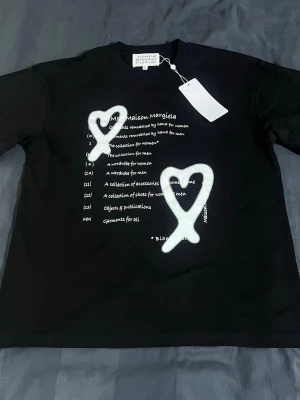 Maison Margiela svart t-shirt med hjärtan - Svart t-shirt från Maison Margiela med vit text och två stora sprayade hjärtan framtill. Klassisk rund hals och korta ärmar. Trycket ger en edgy och unik vibe, perfekt för dig som gillar streetstyle och statement-plagg.