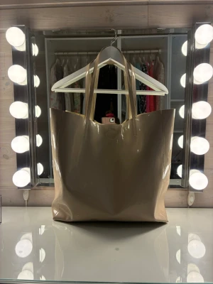 Nude/beige lackad tote bag – rymmer allt (skola/jobb/vardag - Säljer denna super snygga och rymliga tote bag i en jättefin ljusbeige/nude färg med glansig lackfinish! Väskan har en minimalistisk design som passar till allt och rymmer enkelt både laptop, block, sminkväska och andra nödvändigheter. Perfekt som skolväska, till jobbet eller som en stilren vardagsväska.                              Höjd:46 cm, bredd 40 cm, djup: 13 cm. Knäpps med magnet knapp. Väskan har ett innerfack som är 21 cm•13cm som stängs med dragkedja. Mycket fint skick!               