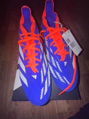 Adidas Predator Pro FG (Nya) - Snygga Adidas Predator fotbollsskor i blått med vita detaljer och orange snörning. Adidas Predator Pro FG i storlek 46. Skorna är helt nya och oanvända, endast uppackade. Originalkartong medföljer.  Perfekta för dig som söker ett par högkvalitativa fotbollsskor med bra grepp och kontroll på naturgräs. Säljs då de inte kommer till användning. Skickar svarar och skickar snabbt. Pris är inte hugget i sten.