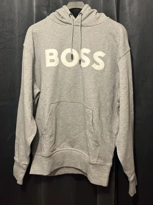 Grå hoodie med BOSS-tryck - Snygg grå hoodie från BOSS med stor vit logga på bröstet. Klassisk känguruficka framtill, ribbade muddar och justerbar huva med dragsko. Tillverkad i mjukt sweatshirtmaterial som är perfekt för en avslappnad stil.