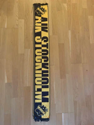 AIK Stockholm halsduk svart/gul - Snygg AIK Stockholm halsduk i svart och gult med klubbens logga på båda sidor. Ena sidan har texten 'AIK STOCKHOLM' och andra sidan 'HATA GÖTEBORG'. Halsduken har fransar i ändarna och är perfekt för matchdagar och supporterliv.
