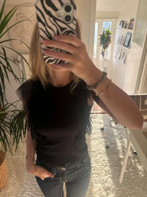 Svart topp med volangdetaljer - Säljer en svart ribbad topp med volangdetaljer i mesh längs axlarna. Toppen är ärmlös och har en rund halsringning. Materialet ser stretchigt och bekvämt ut, perfekt för att lyfta en enkel outfit. Storlek 158/164, Kappahl!!🤍