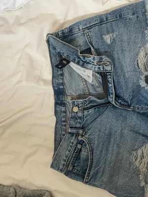 Blå denimshorts från 7S - Säljer ett par klassiska blå denimshorts från 7S med hög midja och slitna detaljer framtill. Shortsen har fem fickor, knappgylf och råa kanter för en avslappnad look. Perfekta till sommaren och lätta att matcha med olika stilar.
